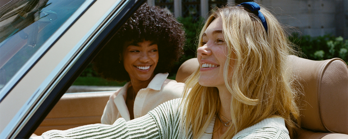 A young white woman smiles while driving a convertible while a young black woman smiles from the passenger seat
