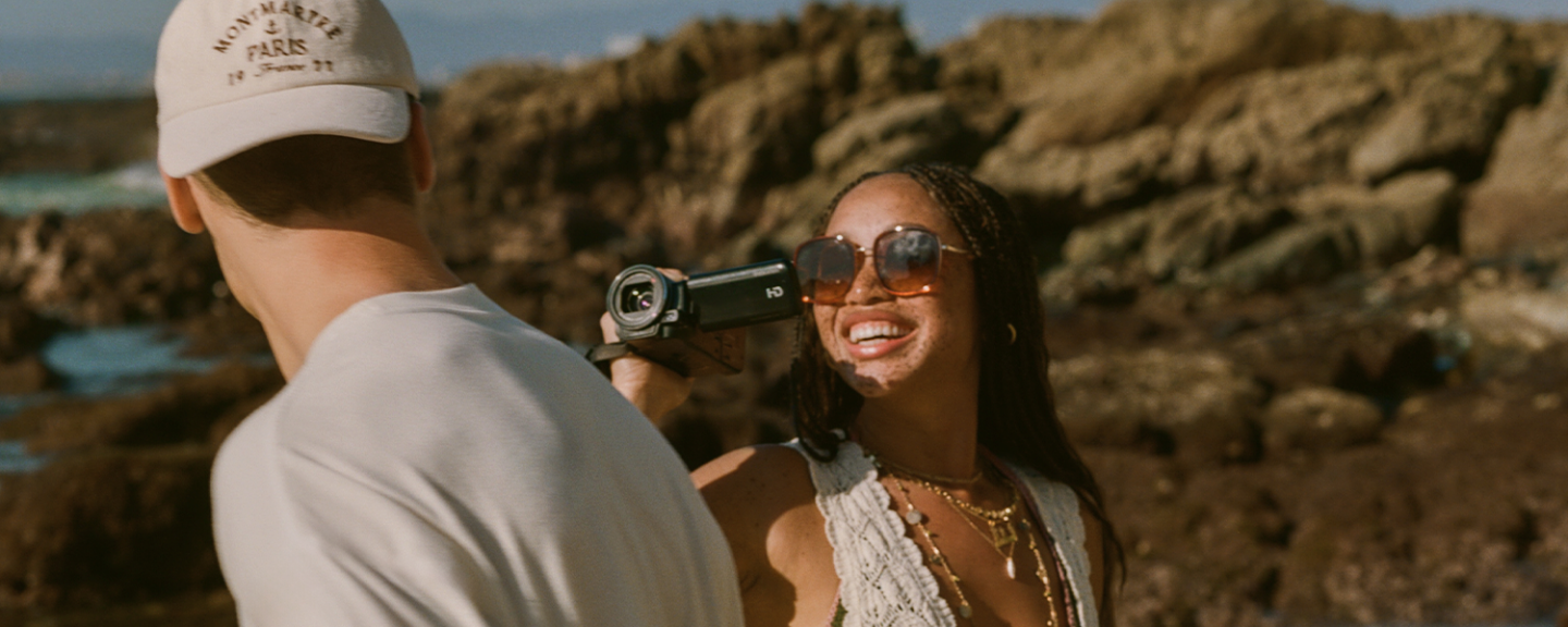 A young black woman smiles while filming a young man at the beach