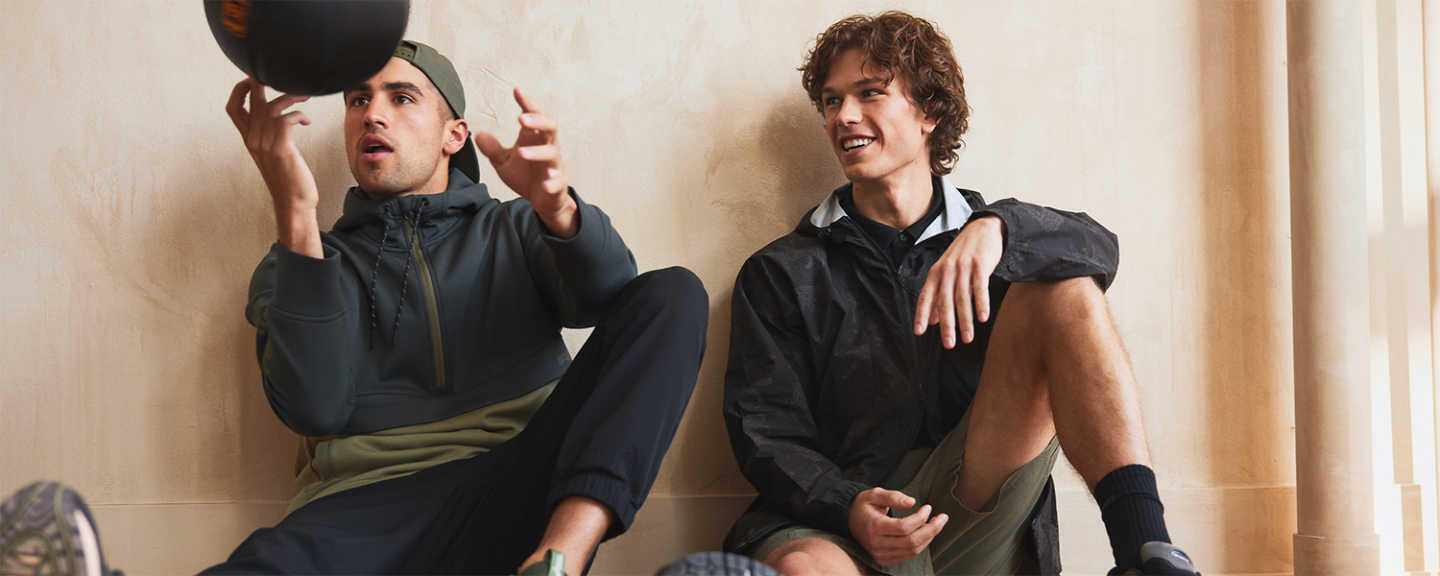 Two young men sit on the floor leaning against a wall, one playing with a basketball