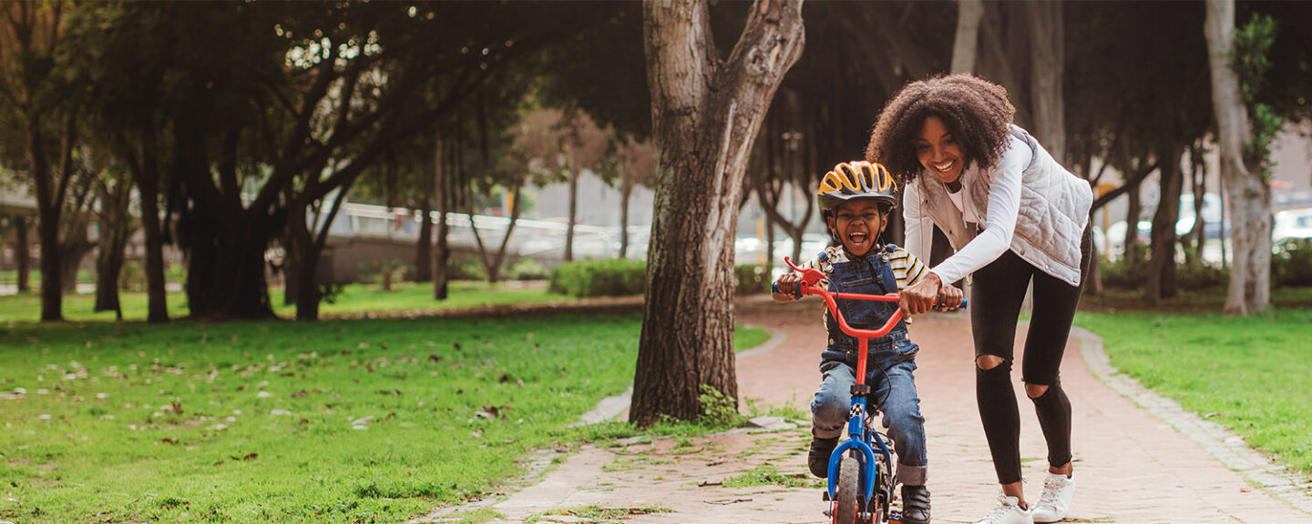 A happy black woman pushes her young son on his bicycle