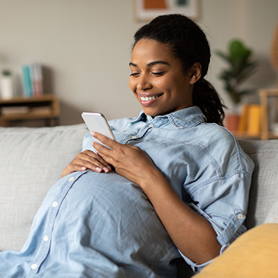 A pregnant black woman smiles while looking at her phone on the couch