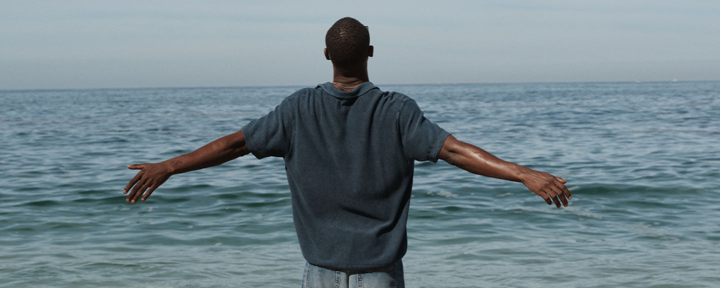 A black man stands on the beach with his arms outstretched