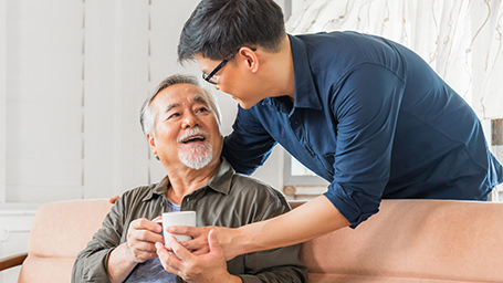 An asian man gives his elderly father a mug of tea